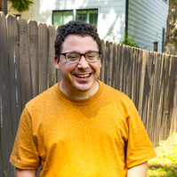 A young man with brown hair and glasses smiles as he stands outside. 