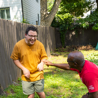 Two men laugh as they stand outside in front of a wooden fence.