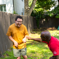 Two men laugh as they stand outside in front of a wooden fence.