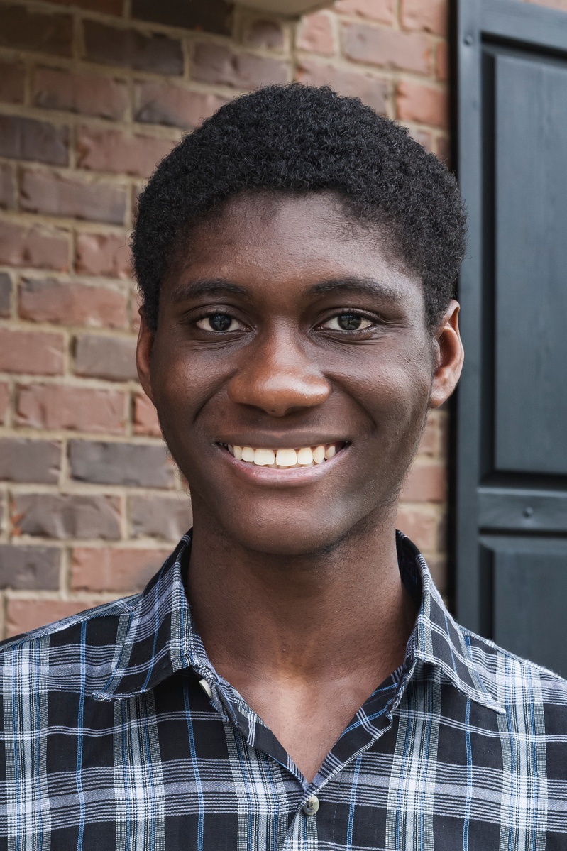 A young African American man in a plaid shirt stands in front of his brick home.