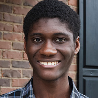 A young African American man in a plaid shirt stands in front of his brick home.