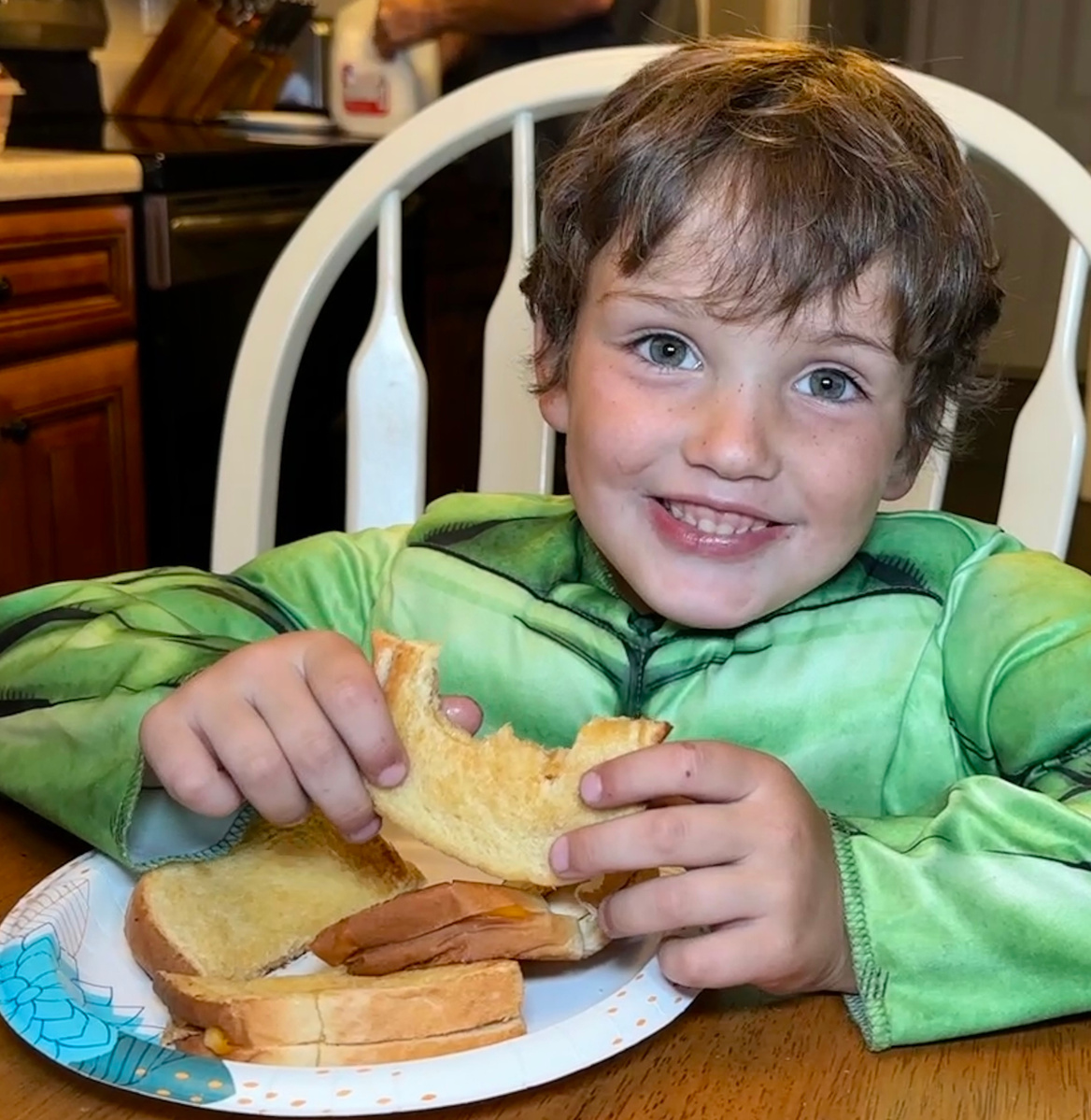 A young boy eats a grilled cheese sandwich while wearing a Hulk costume.