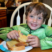 A young boy eats a grilled cheese sandwich while wearing a Hulk costume.