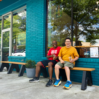 Two men sit on a bench in front of a blue brick store and smile. 