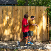 Two men walk next to each other past a wooden fence down a sunny street