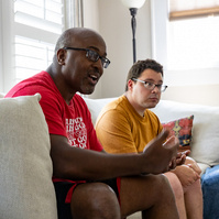 Two men sit on a cream couch in front of sun filled windows. 
