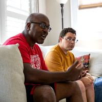 Two men sit on a cream couch in front of sun filled windows. 
