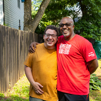 Two men smile as they stand outside in front of a wooden fence.