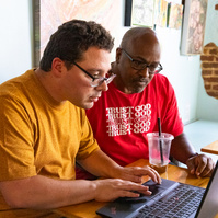 Two men sit next to each other and work on a laptop computer. 