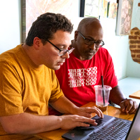 Two men sit next to each other and work on a laptop computer. 
