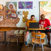 Two men sit next to each other and work on a laptop computer. 