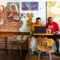 Two men sit next to each other and work on a laptop computer. 