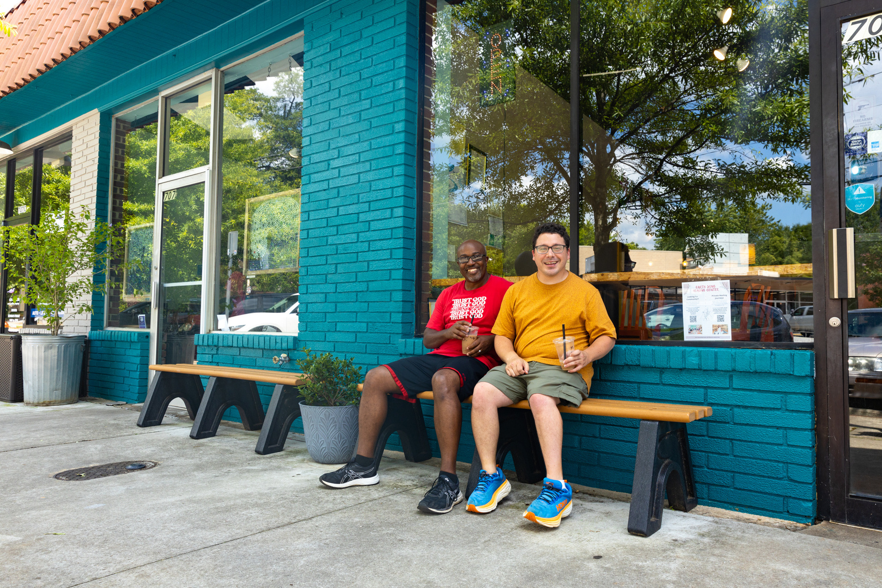 Two men sit on a bench in front of a blue brick store and smile. 