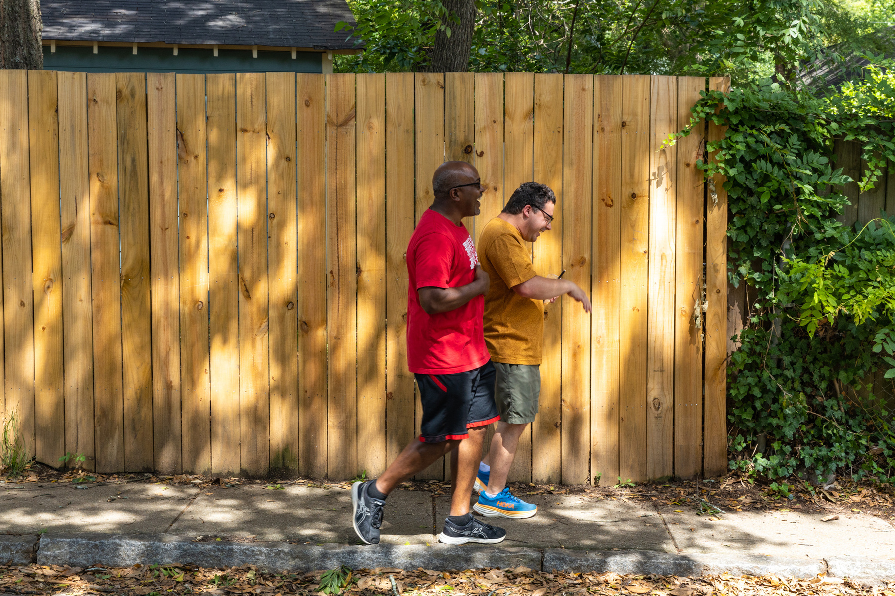 Two men walk next to each other past a wooden fence down a sunny street