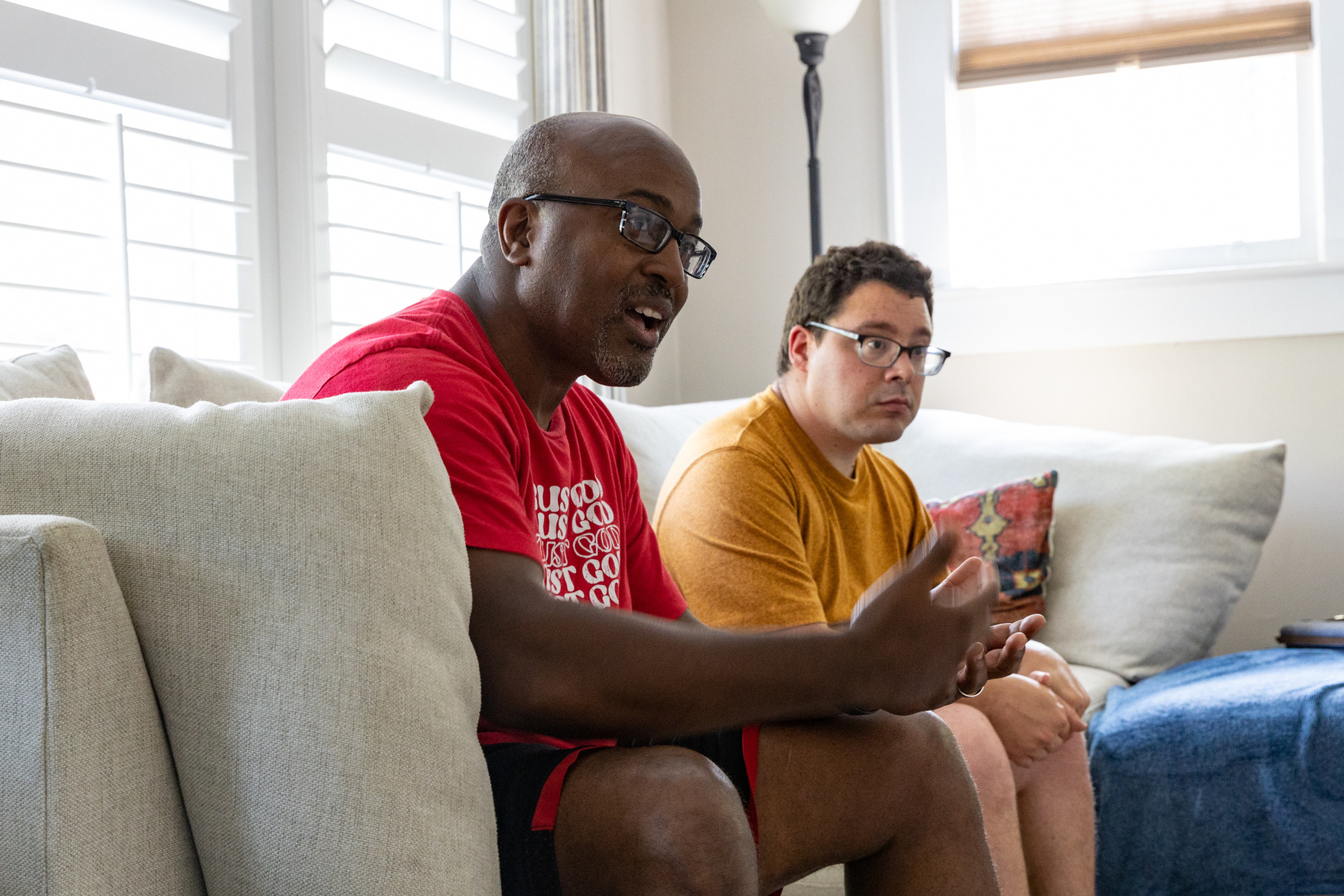 Two men sit on a cream couch in front of sun filled windows. 