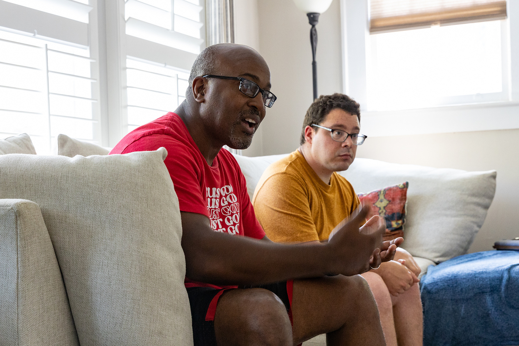 Two men sit on a cream couch in front of sun filled windows. 