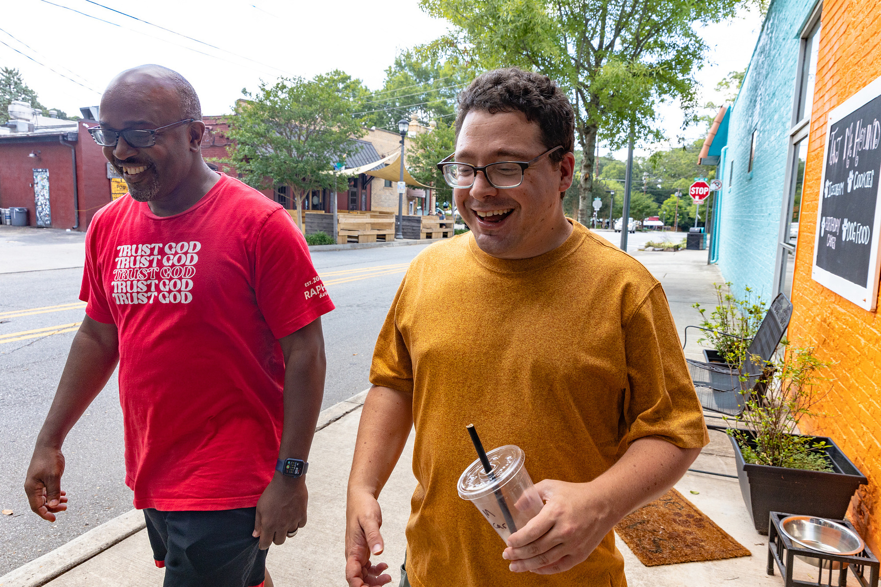 Two men walk next to each other down a sunny street.