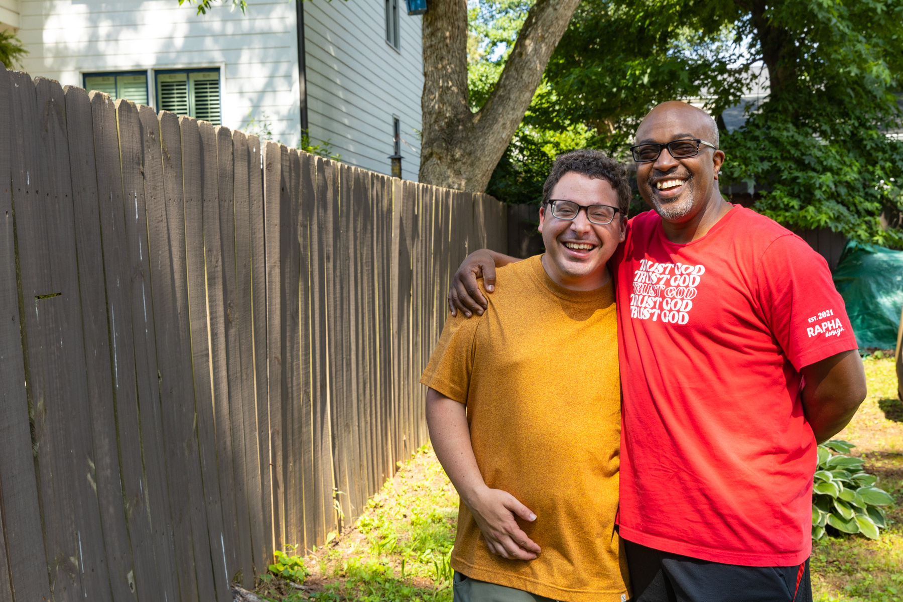 Two men smile as they stand outside in front of a wooden fence.