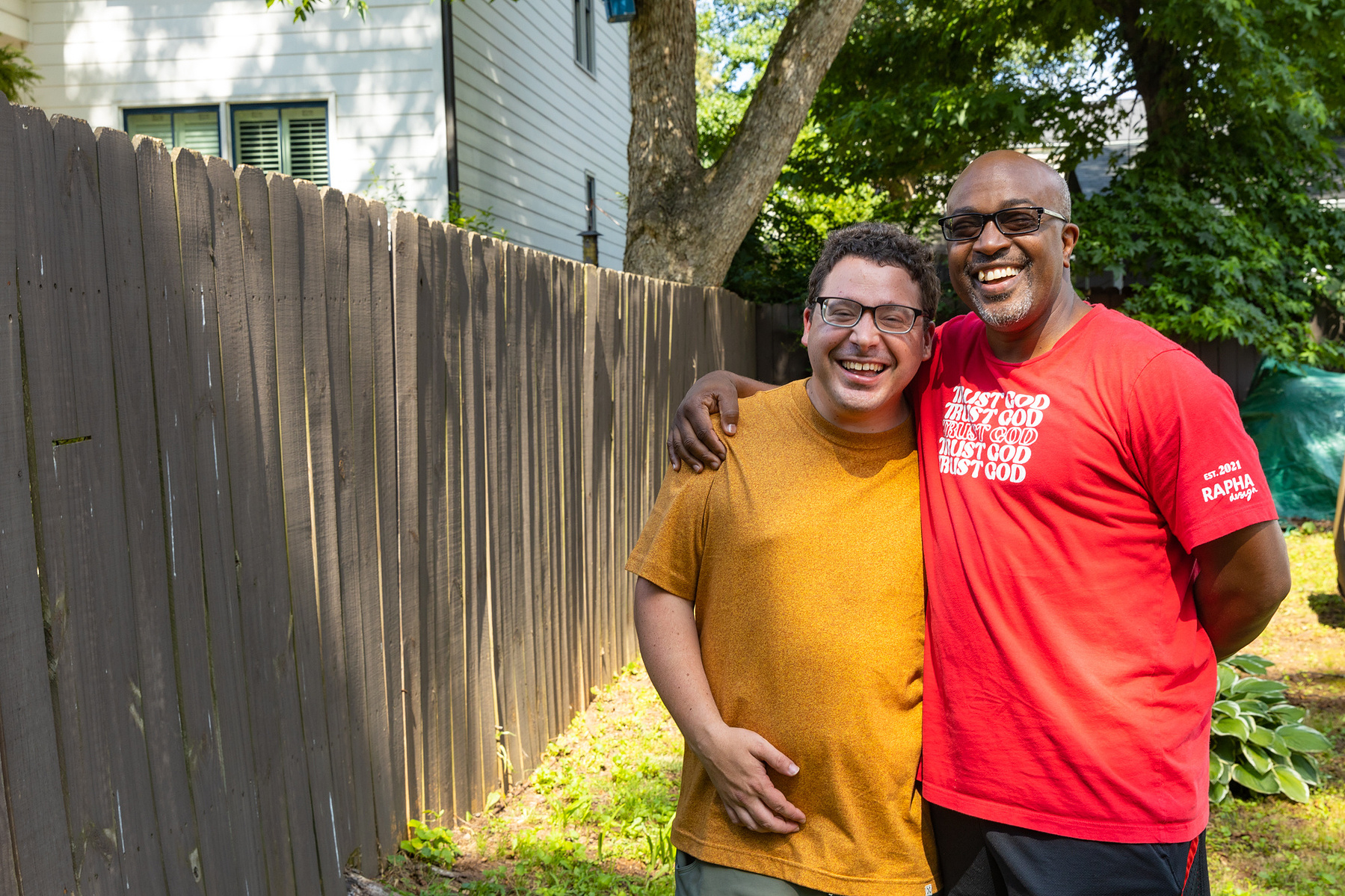 Two men smile as they stand outside in front of a wooden fence.