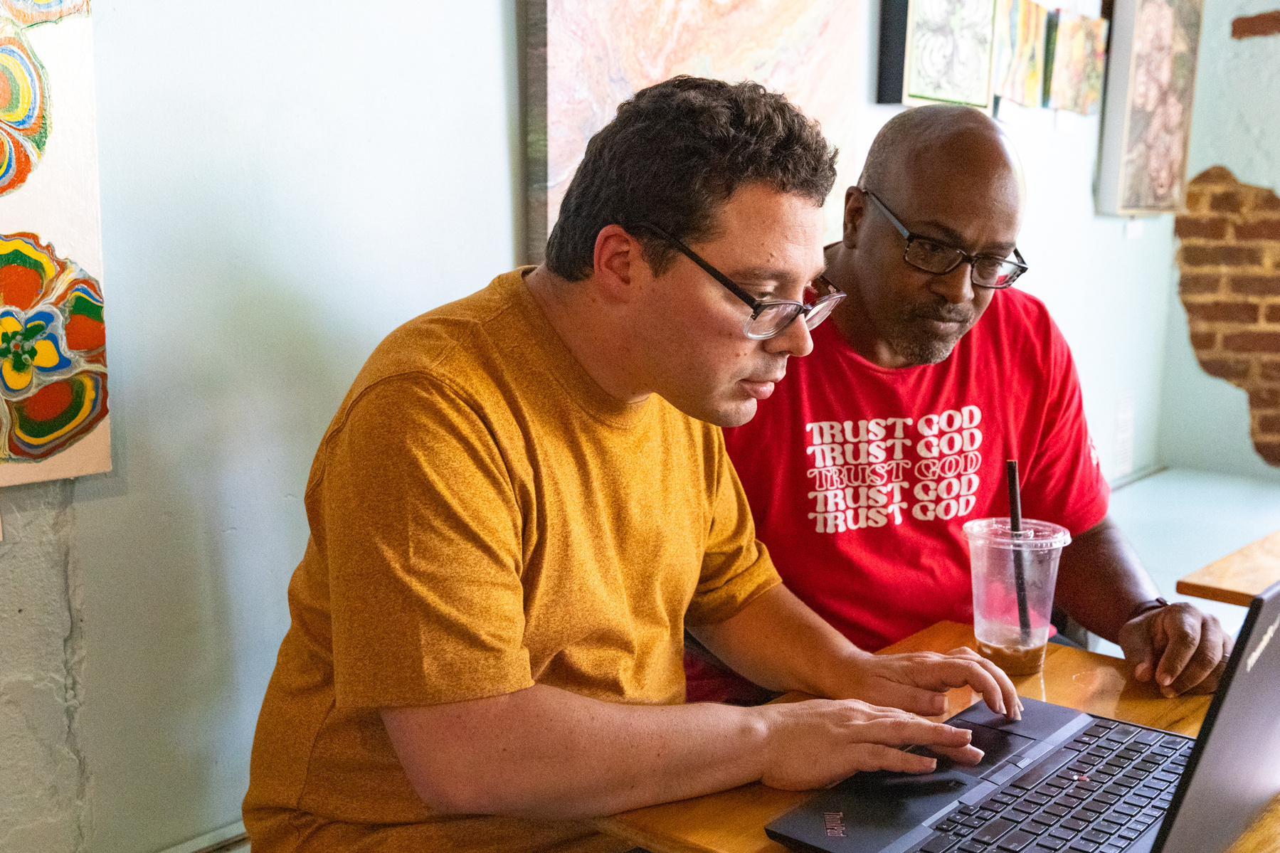 Two men sit next to each other and work on a laptop computer. 