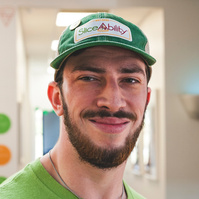 A bearded young man wears a green t-shirt and baseball cap.