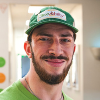 A bearded young man wears a green t-shirt and baseball cap.