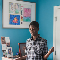 A young African American man in a plaid shirt stands in his blue bedroom. 