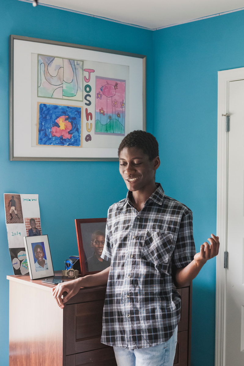 A young African American man in a plaid shirt stands in his blue bedroom. 
