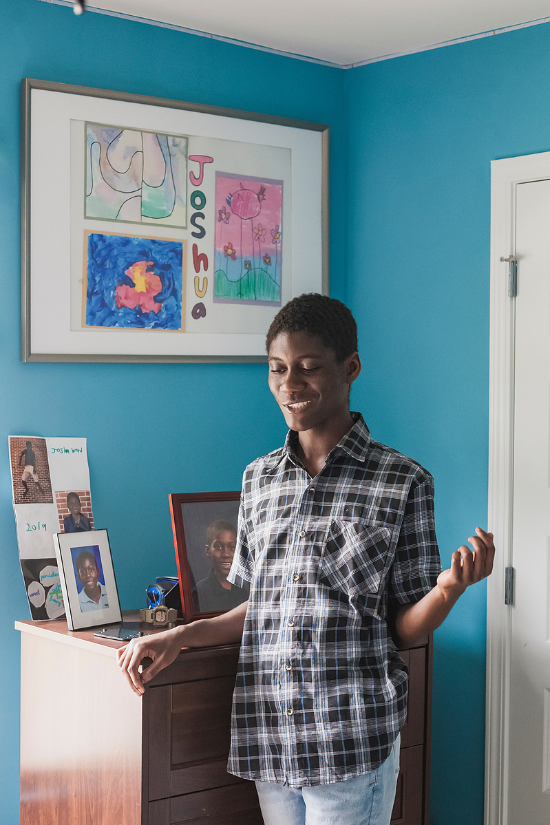 A young African American man in a plaid shirt stands in his blue bedroom. 