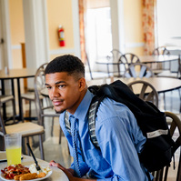 A young African American man wears a blue dress shirt and tie as he dines in a cafeteria.