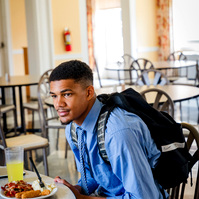 A young African American man wears a blue dress shirt and tie as he dines in a cafeteria.