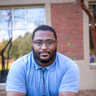 A young black male wears glasses and a light blue polo shirt while standing in front of a brick building.