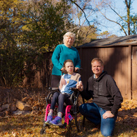 A young girl sits in a wheelchair outside with her grandmother and father surrounding her. 