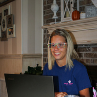 A blond woman wears glasses and sits in front of a laptop computer.  
