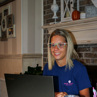 A blond woman wears glasses and sits in front of a laptop computer.  