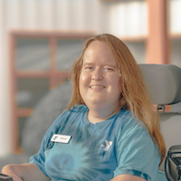 Woman with red hair wears a blue t-shirt and a name tag that reads Caroline. She is sitting in a power wheelchair. 