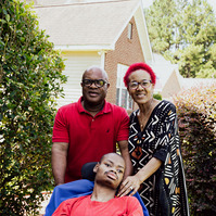 A mother and father stand behind their son as he sits in a wheelchair. 