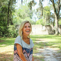 A blond woman in a patterned blouse and jeans stand outside and smiles for the camera. 