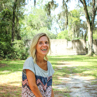 A blond woman in a patterned blouse and jeans stand outside and smiles for the camera. 
