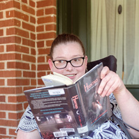 A young brunette woman in a patterned shirt and glasses holds a book and sits on a screened porch. 