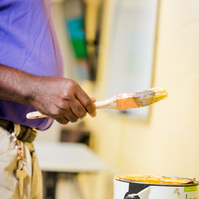 A middle aged African American man wears a purple polo shirt and paints a wall cream.