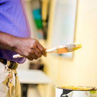A middle aged African American man wears a purple polo shirt and paints a wall cream.