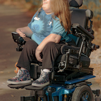 Woman with red hair wears a blue t-shirt is sits in her power wheelchair and smiles.  