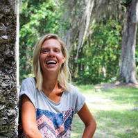 A blond woman in a patterned blouse and jeans stand outside and smiles for the camera. 