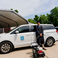 A young African American male wears a cap and gown as he stands in front of a white van and holds a pressure washier. 