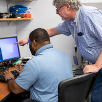 A young man works at a computer while his supervisor assists him on a project. 