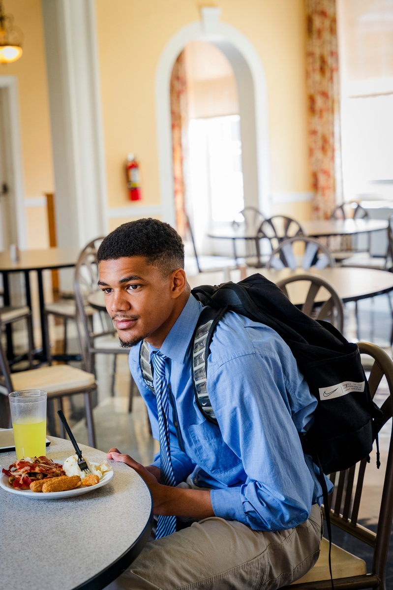 A young African American man wears a blue dress shirt and tie as he dines in a cafeteria.