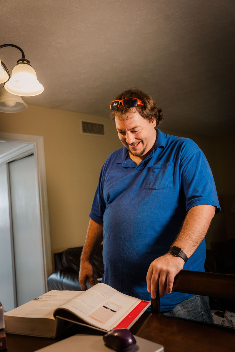 A man stands in a blue polo shirt and looks at a book on a table. 