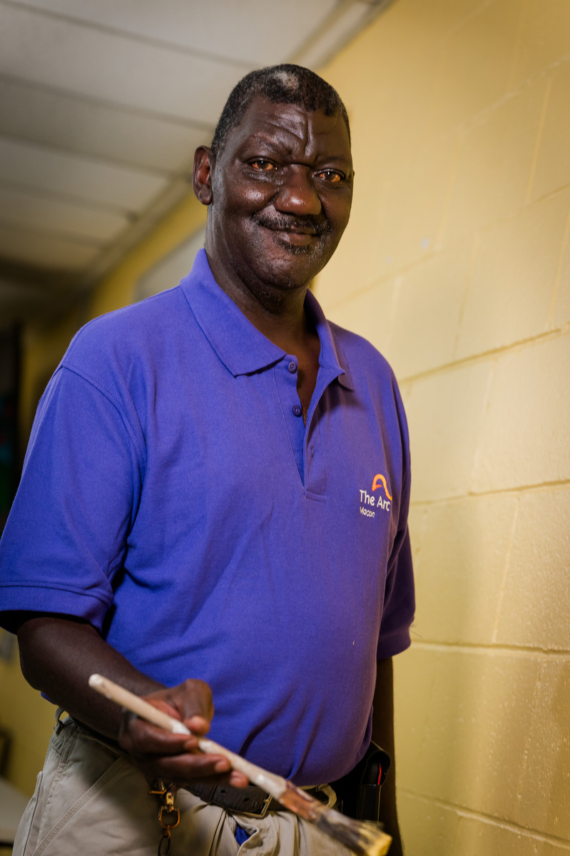 A middle aged African American man wears a purple polo shirt and paints a wall cream.