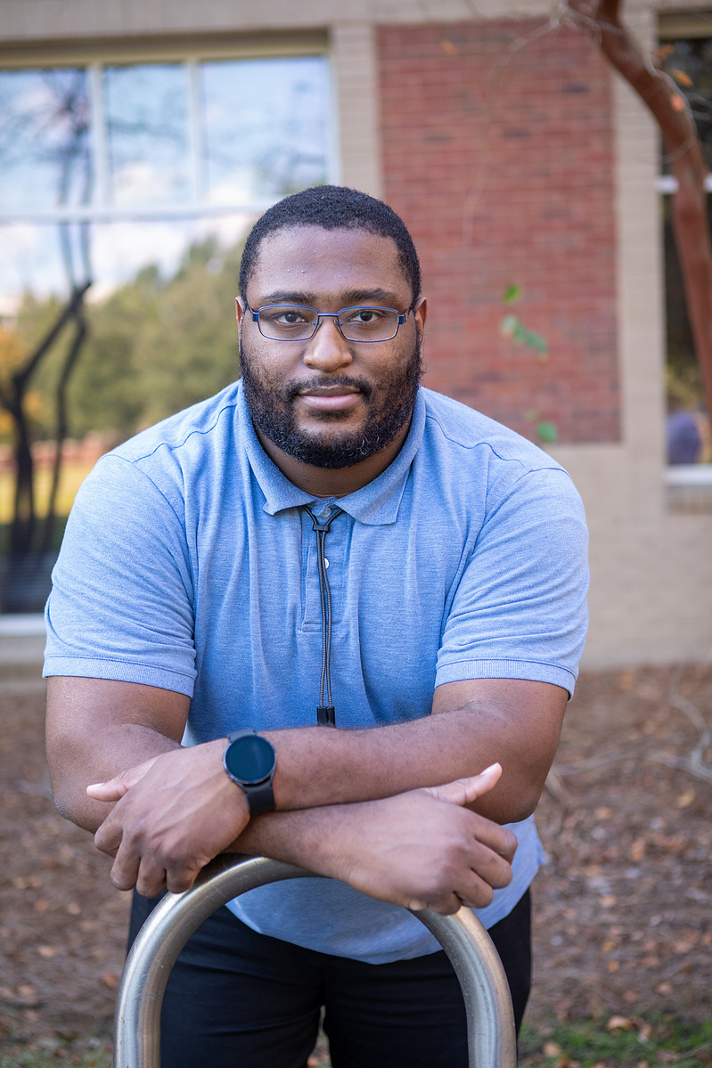 A young black male wears glasses and a light blue polo shirt while standing in front of a brick building.